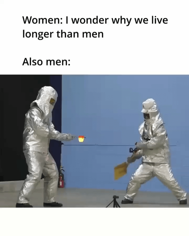 This is a two-panel format meme that uses text and a visual gag to humorously address the stereotype of gender differences in longevity. The top section has text: "Women: I wonder why we live longer than men", followed by "Also men:" above a still image of two men in full silver heat-resistant (firefighter-style) protective suits. One man holds a small container with a burning object, while the other swings a shovel toward the fire in a reckless, playful manner. The joke hinges on the common stereotype that men engage in more dangerous, foolish, or high-risk behaviors that could reduce their life expectancy, directly answering the women's question with absurd visual evidence. The humor comes from the contrast between the extreme protective gear and the careless action that could worsen the small fire.