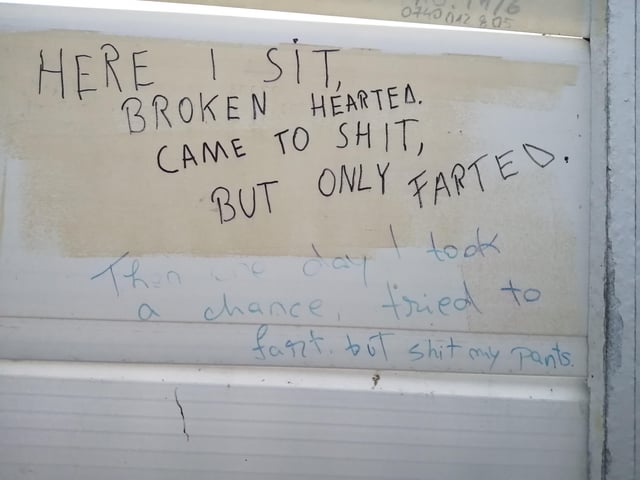 A close-up photograph of a bathroom stall wall featuring handwritten text in casual black and blue ink. The text consists of a two-part humorous poem: the first stanza reads, "HERE I SIT, BROKEN HEARTED. CAME TO SHIT, BUT ONLY FARTED." The second stanza, written in blue ink below, continues, "Then one day I took a chance, tried to fart, but shit my pants." The handwriting is messy with slight smudges, suggesting it was written informally, likely by a college student. The background appears to be a white or off-white stall door with visible lines from the door panels, emphasizing the typical setting of a college bathroom.