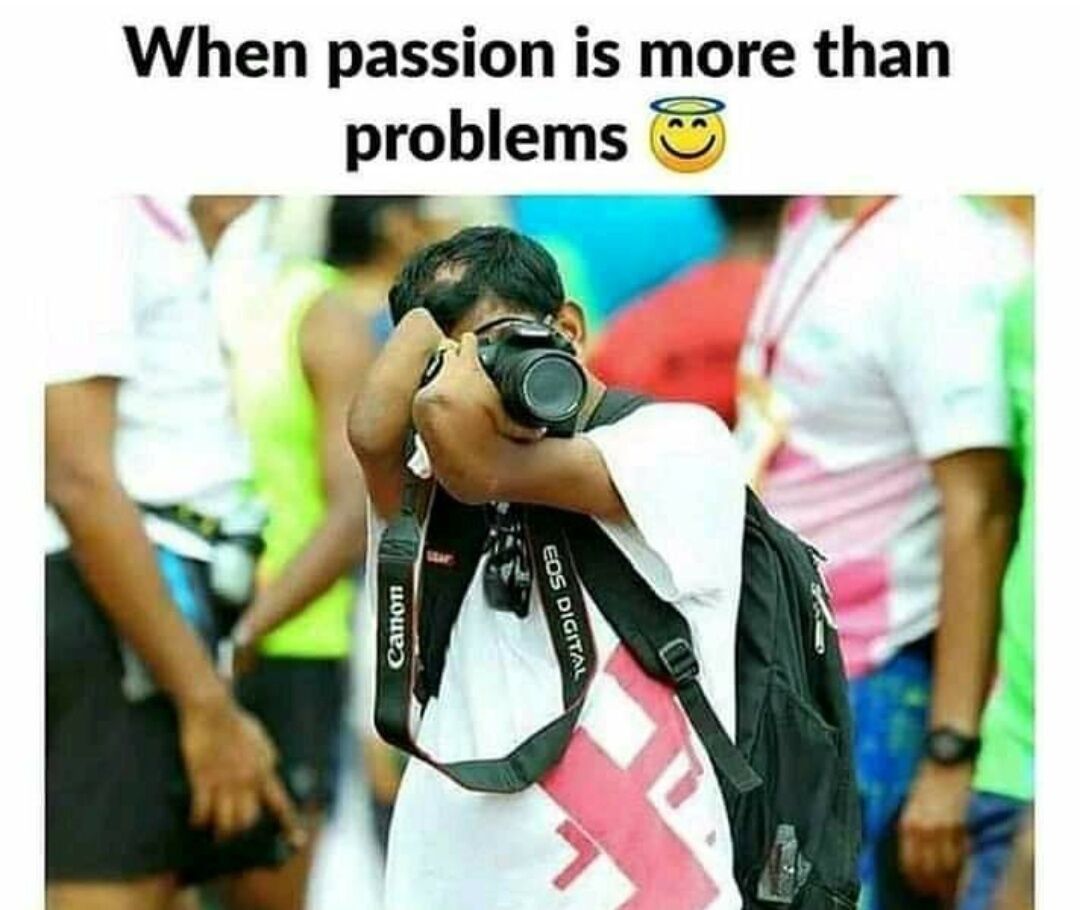 This wholesome meme features a photograph of a man with a physical disability affecting his arms, holding a Canon EOS Digital DSLR camera up to his face to take a photo at a crowded public event. The text overlay at the top reads 'When passion is more than problems' paired with a smiling face with halo emoji, conveying an uplifting message that personal passion can overcome physical challenges and other obstacles. Other attendees are visible blurred in the background of the photo.