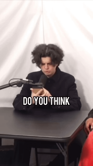 This meme shows a young man with very voluminous, messy dark curly hair sitting at a table during an apparent podcast recording, with microphones positioned in front of him. He is holding a smartphone and looking down at it with a serious, focused expression. Bold white text with a black outline overlaid on the lower part of the image reads 'DO YOU THINK'. The comedic value comes from the unusual, eye-catching shape of the man's hair, which is the reason the original poster states they are sorry for laughing at the clip.