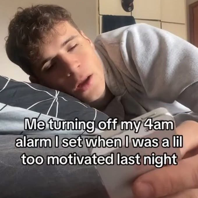 The meme features a young, sleepy man lying face down on a pillow in bed, holding his phone and looking half-awake as if he just turned off an alarm. Overlaid text on the image reads 'Me turning off my 4am alarm I set when I was a lil too motivated last night'. The joke refers to the extremely relatable common experience of feeling overly ambitious about waking up early to be productive the night before, only to be too exhausted and unmotivated to follow through when the alarm rings early in the morning.