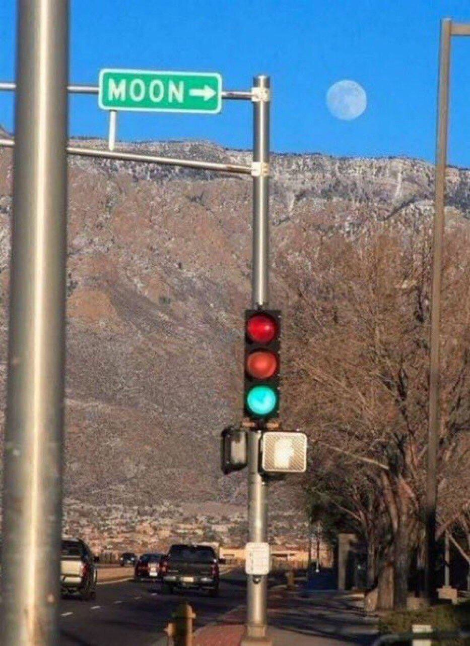 This is an unedited candid photograph of a roadside scene set against a bright clear blue sky, with a rocky, partially snow-dusted mountain range in the background. A green rectangular street sign reading 'MOON' with a right-facing arrow is mounted on a metal pole above a traffic light displaying a green signal. By humorous coincidence, the visible moon in the sky is positioned exactly where the arrow on the street sign points, creating the playful joke that the sign is literally giving directions to travel towards the moon. The scene also includes cars on the road below, bare deciduous trees lining the sidewalk, and no visible people.