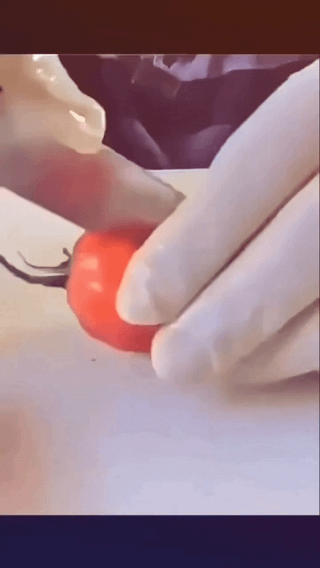This image shows two gloved hands holding a small bright red cherry tomato on a light-colored cutting surface. The positioning of the hands and the tomato creates an accidental suggestive optical illusion that resembles an inappropriate intimate act, which is the source of the meme's humor. The joke plays on the misleading visual that looks sexual at first glance, but is actually just someone handling a tomato.