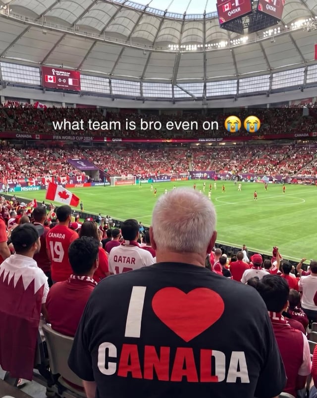 This image is taken from the spectator stands during a 2022 FIFA World Cup match between Canada and Qatar, confirmed by the on-field scoreboards showing a 0-0 tie between the two teams, and surrounding fans wearing Canadian red jerseys or Qatari white and maroon attire. The central focus is an older white-haired man viewed from behind, wearing a black shirt printed with "I ❤️ CANADA". The first letter "C" and final letter "A" of "CANADA" are printed in white, while the middle four letters are printed in red, creating a visual trick that makes the red portion of the text appear to spell "QATAR". Overlaid at the top of the image is white text that reads "what team is bro even on" followed by two crying laughing emojis, mocking the ambiguous dual-team reference on the fan's shirt.