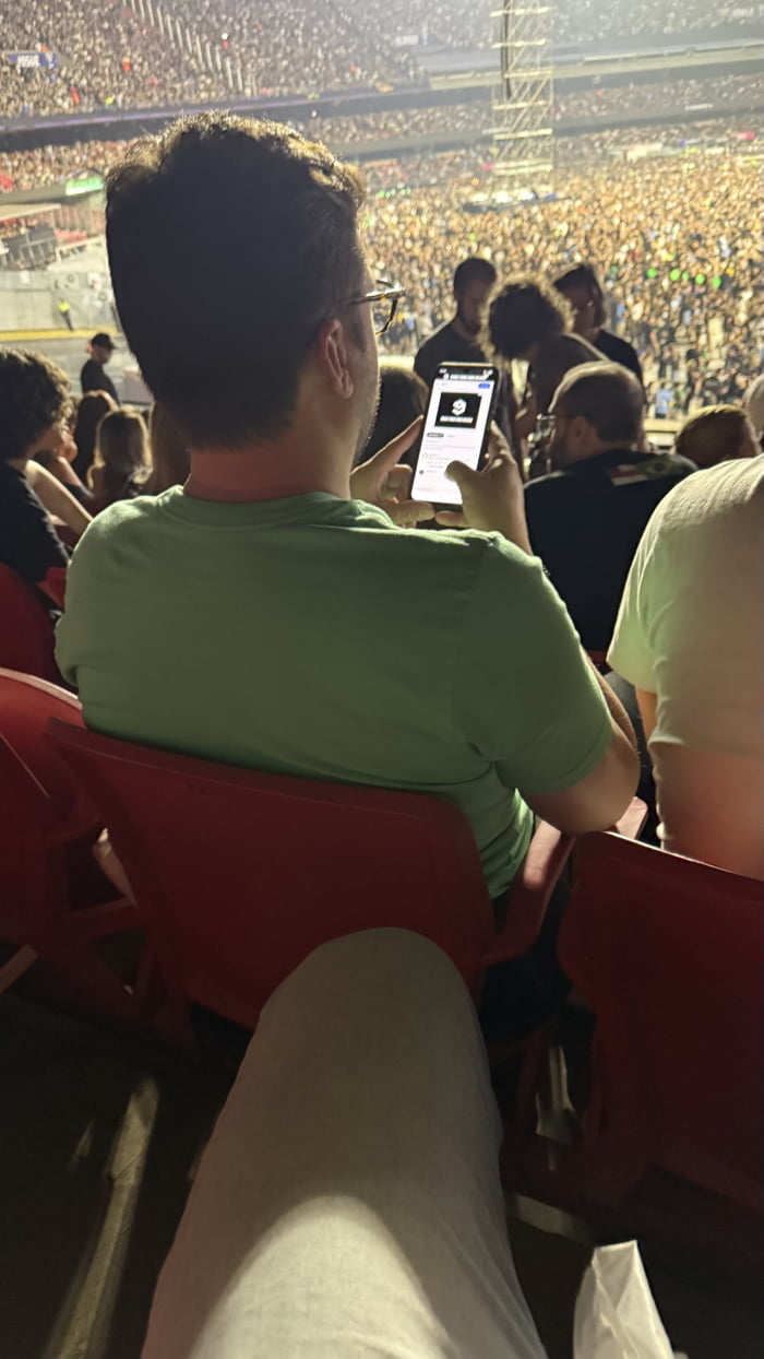 A photograph taken from behind a person wearing a green shirt seated in red stadium seats. The individual is holding a smartphone in their hands, possibly interacting with the device, while surrounded by a large, densely packed crowd of spectators. The stadium is brightly lit, with the audience extending to the far end, indicating a major live event (concert, sports match, etc.). The perspective emphasizes the contrast between the person's focus on their phone and the surrounding engagement with the live environment.