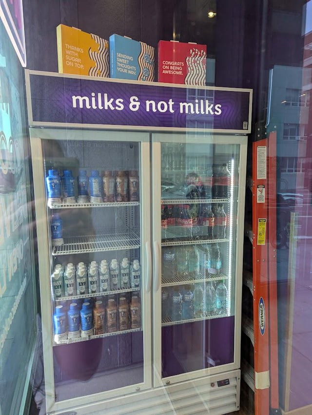 The image is a candid photograph of a double-door glass-front commercial beverage refrigerator inside a retail store. Above the fridge, a purple sign reads 'milks & not milks', a humorously broad, logically exhaustive categorization that splits all possible contents into two groups: items that are milk, and all items that are not milk. The left section of the fridge holds various bottled milk and coffee-milk beverages, while the right section holds sodas, bottled water, and other non-milk drinks. The joke comes from the fact that this classification is technically correct and literally covers every possible object in existence, which aligns with the meme title stating the fridge must contain everything in the universe by definition.