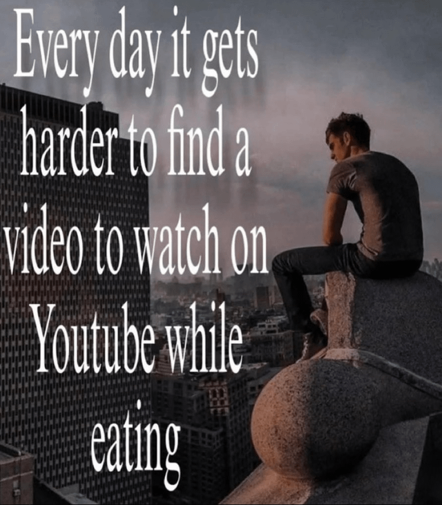 The image features a man sitting on a stone architectural ledge of a tall building, overlooking a dense urban cityscape under a cloudy sky. He is positioned with his back to the viewer, wearing a gray t-shirt and dark pants, legs hanging over the edge, and appears contemplative. To the left of the man, large white serif text reads: 'Every day it gets harder to find a video to watch on Youtube while eating'. The scene combines a somber, reflective visual tone with humorous, relatable text about the everyday frustration of finding engaging content during meals.