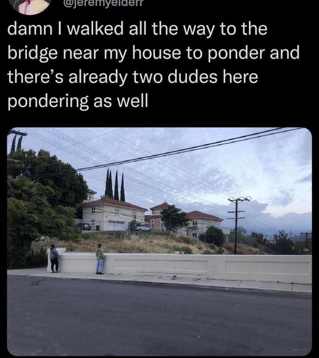 This is a screenshot of a social media post, consisting of text at the top and a photograph below. The text laments that the poster walked to a local bridge to have private time to think deeply, only to find two other men already at the same bridge doing the same activity. The accompanying photo shows a low concrete roadside barrier overlooking a residential hillside neighborhood, with two men standing facing away from the camera, leaning against the barrier and appearing to be lost in thought as they look out at the view. The humor comes from the relatable, unspoken shared experience of seeking out a quiet private spot to reflect, only to discover other people had the exact same idea.