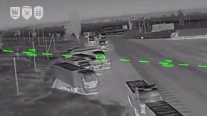 The image is a monochrome thermal drone feed capturing a line of multiple military supply trucks parked along a paved road in an open rural area. Bright green digital overlays including targeting reticles and text reading 'FIRE POINT' are marked on and near the vehicles, designating them as active targets for an incoming strike. Three official military unit insignia are displayed in the upper left corner of the footage.