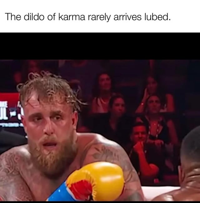 A meme featuring a top text overlay that reads 'The dildo of karma rarely arrives lubed' above a photograph of an intense, sweaty male boxer in a boxing ring. The boxer has visible tattoos, disheveled hair, and is wearing yellow and blue gloves while facing another opponent. The background shows a blurred audience watching the match, with some spectators appearing engaged. The text uses a sexual innuendo ('dildo', 'lubed') to humorously convey that karma (consequences) typically arrives harshly or without ease.