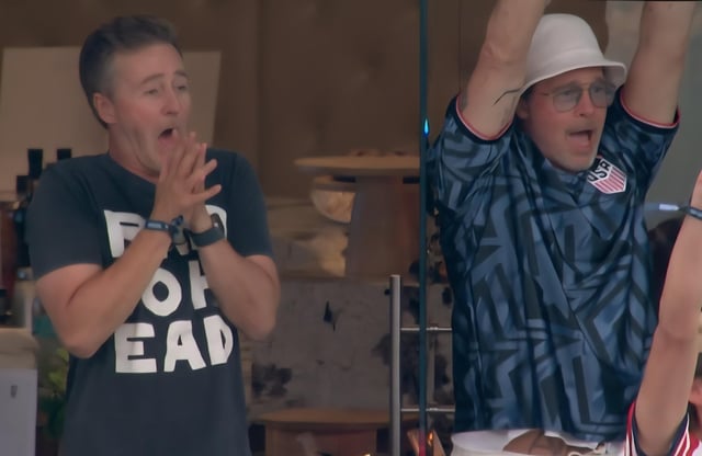 The image initially appears to show two separate excited men, seemingly watching a sports event. The figure on the left wears a dark t-shirt with large white text, clasps his hands near his open mouth in a shocked excited expression, and wears two wrist accessories. The figure on the right wears a white bucket hat, glasses, a blue geometric patterned US soccer jersey, and holds both arms raised above his head cheering. The image relies on an optical illusion: a clear glass panel sits between the viewer and the man on the right, so the 'second man' on the left is actually just his reflection on the glass, meaning only one real person is present in the scene.