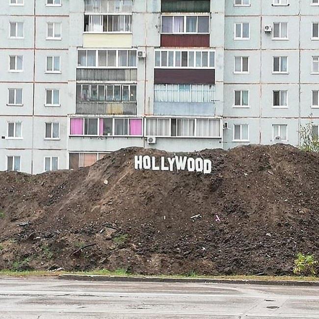 The image depicts a large, unattractive pile of construction dirt positioned in the foreground of a drab, grey, multi-story Soviet-era prefabricated apartment building. White block letters spelling 'HOLLYWOOD' are placed along the crest of the dirt hill, directly mimicking the iconic Hollywood Sign located in the Hollywood Hills of Los Angeles. The intentionally absurd juxtaposition between the glamorous, world-famous Hollywood landmark and the mundane, unglamorous setting of a run-down urban construction site creates a sarcastic, humorous effect.