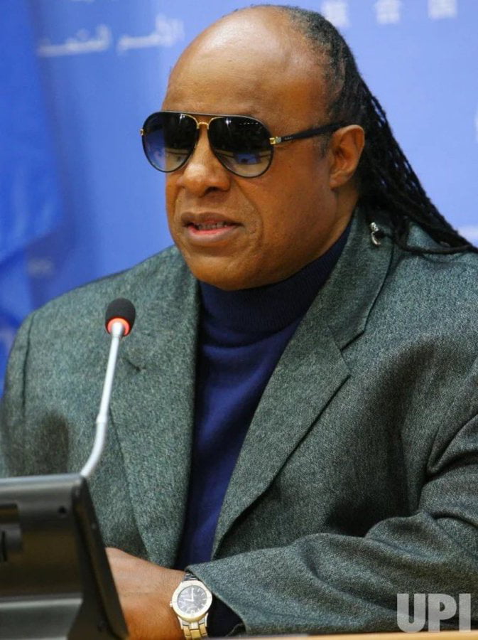 A photograph of Stevie Wonder at what appears to be a press conference or formal event. He is seated at a table, wearing a gray blazer over a blue turtleneck, dark sunglasses, and a wristwatch on his left wrist. A microphone is positioned in front of him, and part of a laptop is visible on the table. The image is accompanied by the text question: 'Can someone explain why Stevie Wonder wears a watch?', which plays on the irony of a blind individual wearing a watch, though in reality, visually impaired individuals often use talking watches or tactile timepieces.