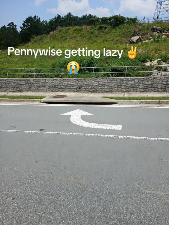 A meme image featuring a photograph of a road with a white directional arrow painted on the asphalt, pointing upward and curving slightly to the right. In the background, there is a grassy embankment with a stone retaining wall and a metal fence. Overlaid at the top of the image is the text 'Pennywise getting lazy' accompanied by a yellow laughing emoji (🤣) and a peace sign emoji (✌️). The humor derives from the road arrow resembling the iconic upward-pointing hand gesture of Pennywise the Dancing Clown from Stephen King's 'It,' presented here as a 'lazy' version since it is merely a painted road marking rather than the character's dramatic pose.