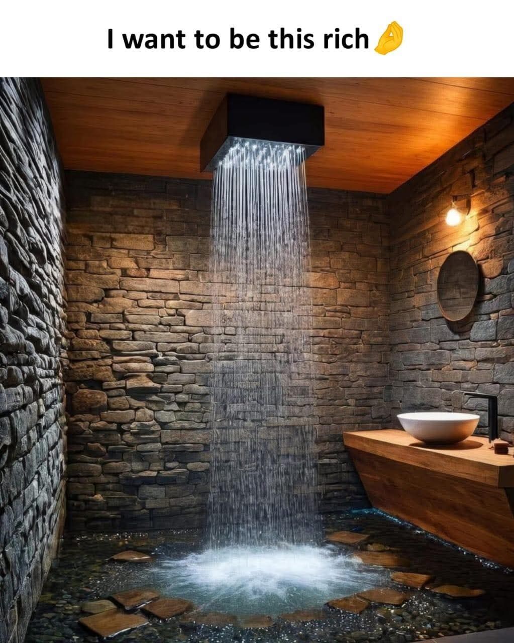 This is a relatable aspiration meme. The bottom portion of the image displays an extremely opulent custom bathroom: it has rough stacked natural stone walls, a warm wooden ceiling, a large square overhead waterfall showerhead pouring a heavy stream of water down into an open shower area with a pebble-covered floor and flat stone stepping stones. To the right, there is a modern wooden vanity with a round vessel sink and a small round wall mirror. Above this image, black text reads 'I want to be this rich' next to a yellow folded hands prayer emoji, expressing the common desire to have enough wealth to afford such a high-end, luxury home feature.