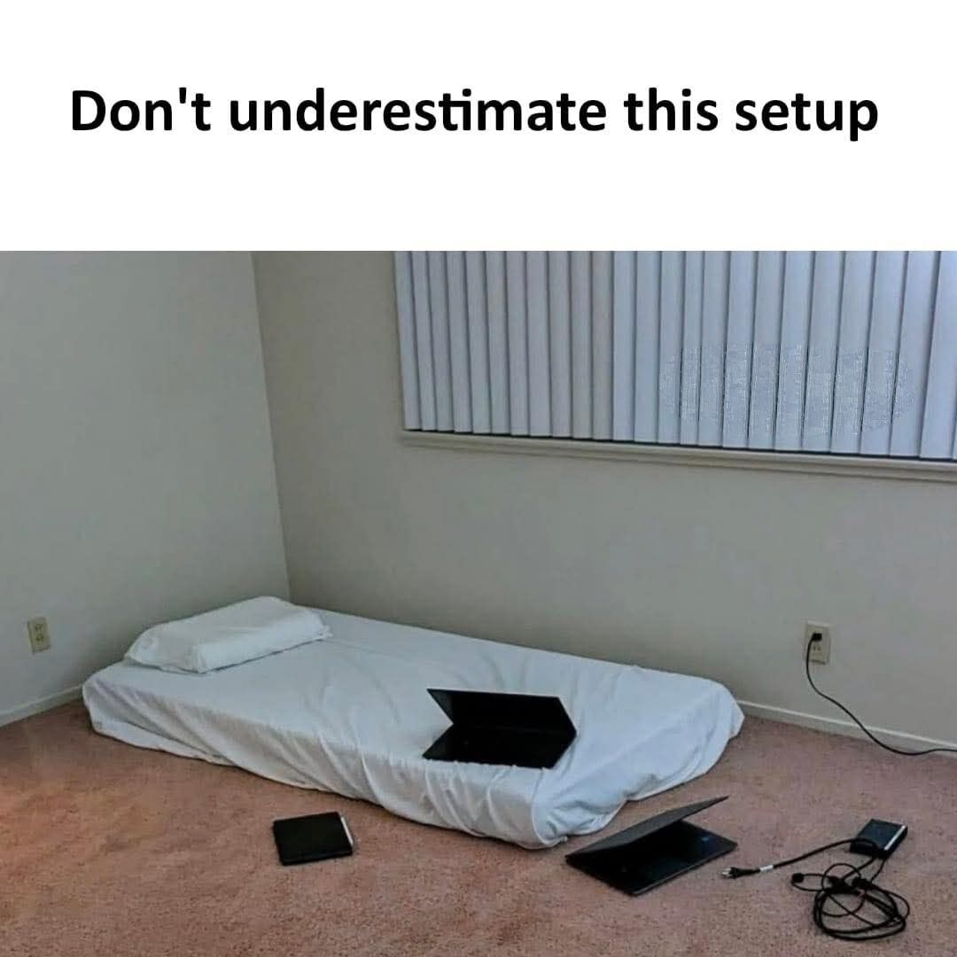 The meme features bold black text at the top reading 'Don't underestimate this setup' positioned above a photograph of a sparse, unfurnished bedroom corner. The scene shows a low twin mattress placed directly on light pink carpet with no bed frame, fitted with a white sheet and a single white pillow. One black laptop sits open on the mattress, while two additional open laptops, a closed tablet, a charging brick, and loose cables are scattered on the carpet next to the mattress. Vertical white blinds cover the only window on the plain light gray wall. The joke relies on the contrast between the seemingly unimpressive, basic living space and the implication that the setup is highly functional for remote work, content creation, or gaming, resonating with people who prioritize utility over expensive home decor.