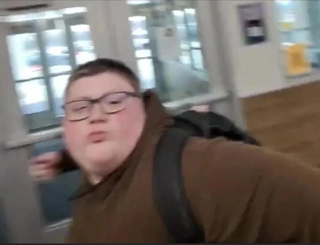 This image captures a young, heavyset boy wearing glasses, a brown hoodie, and a black backpack, making an exaggerated pouty, pursed-lip facial expression. He is positioned in an indoor public space, likely a school or building entryway, with glass doors visible in the background. His body is slightly turned, and one arm is raised in mid-motion, which amplifies the comedic, over-the-top nature of his annoyed look. This image is widely used as a reaction meme online to convey feelings of petulance, playful frustration, or exaggerated annoyance in response to frustrating situations, petty comments, or minor inconveniences.