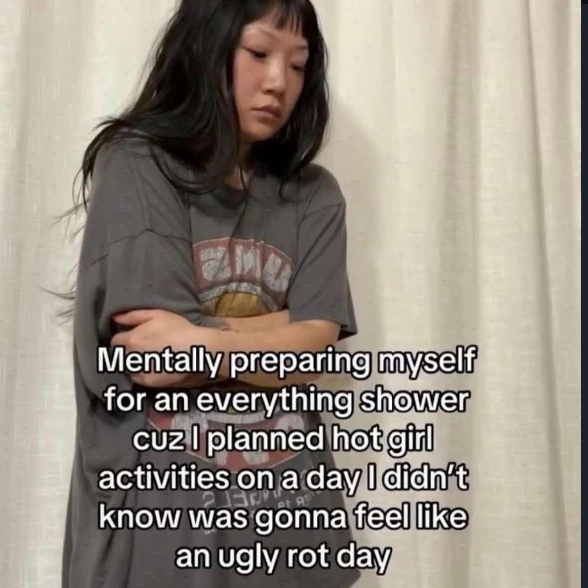 The meme features a young East Asian woman with long dark hair and blunt bangs wearing an oversized gray graphic t-shirt, standing in front of a light beige curtain with her arms crossed across her chest. She has a tired, unmotivated, apathetic expression as she looks downward. White overlaid text on the image reads: 'Mentally preparing myself for an everything shower cuz I planned hot girl activities on a day I didn't know was gonna feel like an ugly rot day'. The joke plays on the common relatable experience of having to force oneself to complete an extensive full-body grooming routine (referred to as an 'everything shower' in slang) to follow through with planned fun outings or self-care plans, when one unexpectedly feels too exhausted and lazy to leave the house on what is called a 'rot day' online.