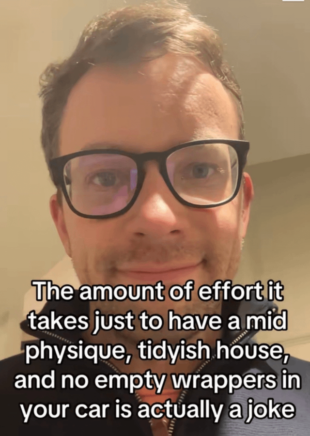 A meme image featuring a real human male with short, slightly messy hair and black-framed glasses, standing indoors against a plain wall with a neutral expression and a faint smile. Overlaid in bold white text across the lower half of the image is a humorous complaint: 'The amount of effort it takes just to have a mid physique, tidyish house, and no empty wrappers in your car is actually a joke.' The image humorously captures the relatable struggle of maintaining basic adult responsibilities that are often dismissed as 'easy' or 'light work.'