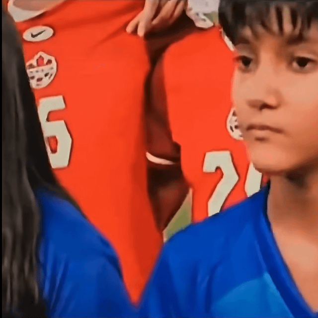 This meme uses a forced perspective optical illusion for humorous effect. In the foreground, part of a girl with long dark hair wearing a blue sports top is visible on the left, and a young boy in a blue sports jersey with a neutral, slightly serious expression looking off-camera is on the right. In the background, people wearing red sports jerseys (one with the number 6 and a Nike logo) are present: a hand resting on the back of the person in the red number 6 jersey is positioned such that from the camera angle, it appears to be placed on the chest of the girl in the foreground. The title references the joke that if the girl had noticed this misleading apparent placement, she would likely have had a negative reaction, making it 'good she didn't overreact' to the illusion.
