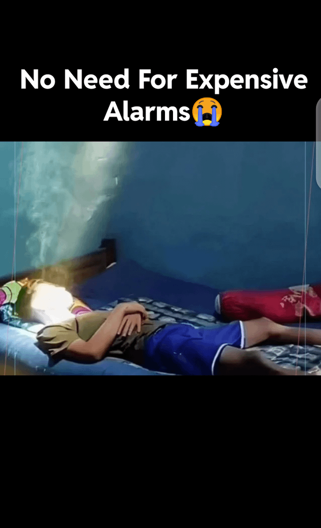 This humorous meme features a candid photo of a young person lying asleep on their back on a bed in a small room. A bright, sharp ray of morning sunlight is shining directly onto the sleeping person's face, which is framed as a free alternative to purchasing expensive alarm clocks. The top text paired with a crying emoji jokes that this natural, unavoidable bright light is highly effective at waking people up, even if it is an unpleasant way to start the day, making the content relatable to anyone who has had their sleep interrupted by unplanned bright morning sun through their window.