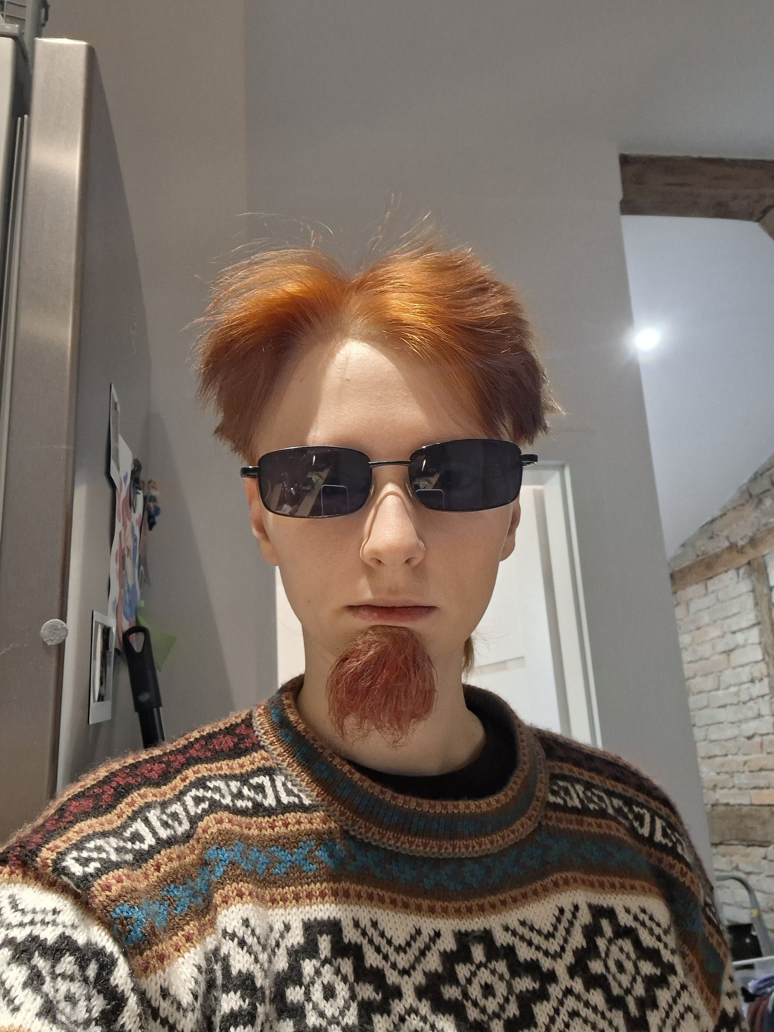 A portrait of a person with red spiky hair and a small beard (soul patch) wearing black rectangular sunglasses. They are dressed in a patterned sweater featuring colors including brown, blue, white, and black with intricate geometric designs. The background shows an indoor setting, likely a home, with a stainless steel refrigerator on the left, wall decorations, a white door, and a section of exposed brick wall on the right. The individual is standing still and facing the camera with a neutral, serious expression.