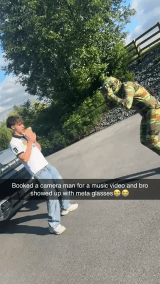 The image is an outdoor scene set on an asphalt road next to green foliage and a wooden fence. A young man in a white polo shirt and light blue jeans leans against a car holding a drink, posing as if for a music video shoot. Across from him, a person wearing a full green and brown camouflage ghillie suit holds their hands up in a pose mimicking someone operating a professional camera, pretending to film. A text overlay at the bottom jokes that the booked cameraman arrived to the shoot with only Meta smart glasses as his filming equipment instead of professional gear. The humor is twofold: the absurdity of the cameraman wearing full camouflage for a standard music video shoot, and the joke that he is using consumer-grade smart glasses to film instead of proper camera equipment. The original post title adds a second layer of humor, noting that the client paid extra money specifically for the camouflage feature of the cameraman's service.