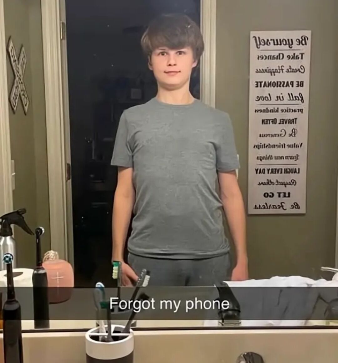 This is a humorous single-panel photo meme set in a home bathroom. A young man stands in front of a bathroom mirror posed as if taking a mirror selfie. An overlaid text caption at the bottom reads "Forgot my phone", creating an ironic joke: the subject is clearly posed for a selfie but claims to have forgotten the device needed to take one, implying the photo was taken with a different device or leaning into the absurdity of the selfie pose without a phone present. A secondary minor humorous detail is the reversed motivational quote wall sign visible in the mirror reflection on the right side of the frame. Basic toiletries including toothbrushes, a spray bottle, and a soap dispenser are visible on the bathroom counter in the foreground.