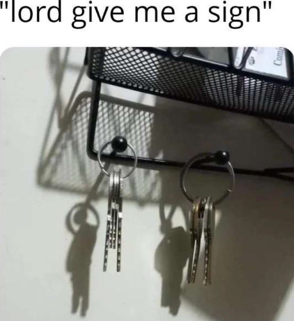 The meme features a line of text at the top that reads 'lord give me a sign', placed above a photograph of two key rings with multiple keys hanging from hooks on a black mesh desk organizer. The shadows cast by the key rings and keys onto the flat white surface below coincidentally form the distinct shapes of two human figures, each appearing to carry a backpack or shoulder bag while walking away. The humor comes from the visual pun where the accidental shadow formation acts as the 'sign' the speaker asked for from a higher power.