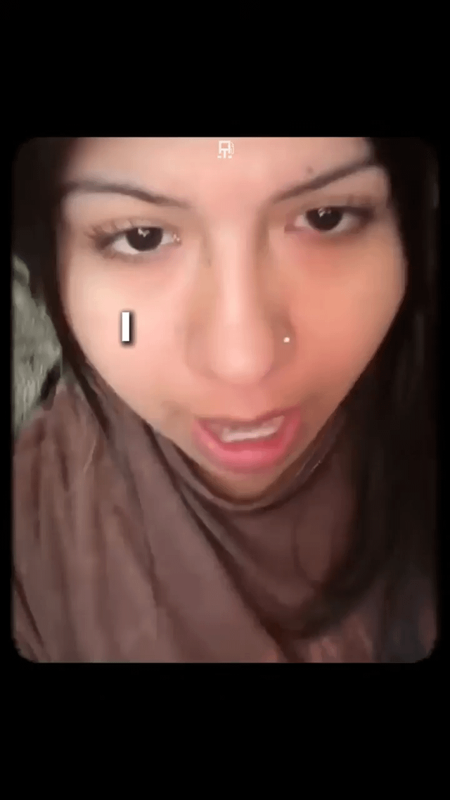 This meme is a close-up shot of a young woman with dark hair, a small nose stud piercing, and wearing a brown turtleneck garment. She has a visibly annoyed, deadpan, unamused expression as she stares directly at the camera. A small white vertical loading progress bar is edited onto her left cheek, creating a visual gag that implies she is buffering, processing, or reacting with exasperation to whatever event the title 'She asked for it' refers to.