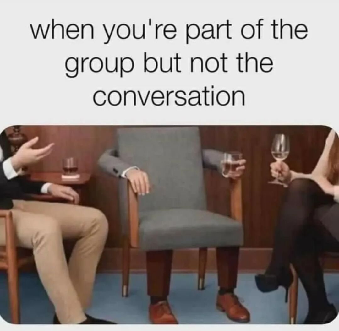 The meme features text at the top stating 'when you're part of the group but not the conversation' placed above a surreal edited photograph. The photograph shows three people sitting in a row during what appears to be a casual social gathering with drinks. The middle individual's head and torso are replaced by a grey upholstered armchair, with only their arms, legs, and dress shoes visible, holding a drink just like the other two people on either side. The image humorously personifies the common relatable feeling of being physically present in a social group but being excluded from or unable to participate in the ongoing conversation, feeling as unnoticeable and inanimate as a piece of furniture.