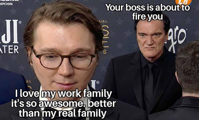 A meme featuring two real people in a formal setting (likely a red carpet or event, indicated by a starry backdrop and suits). The foreground person expresses sincere affection for their 'work family,' claiming it is 'so awesome' and 'better than my real family.' In contrast, the background person (presumed to be their boss) has an overlay text stating 'Your boss is about to fire you,' creating an ironic contrast between the person's praise of their work family and the imminent job loss.