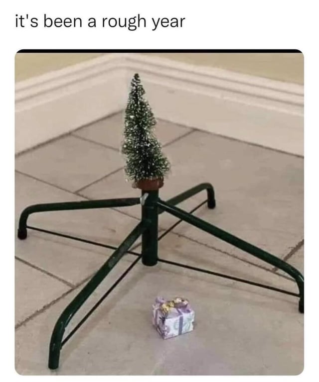 The meme features a photograph of a minimalist Christmas setup. A small, sparse artificial Christmas tree (approximately 6-8 inches tall) is placed directly on a green metal tree stand, with no full-sized tree attached. Next to the stand on the tiled floor lies a tiny purple gift box with a gold bow. The background shows a white baseboard along the wall. Above the image, the text reads 'it's been a rough year,' creating a humorous and relatable commentary on having a difficult year, reflected in the underwhelming, scaled-down holiday decorations.