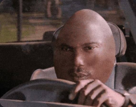 This is an absurd edited meme featuring a heavily distorted version of Kanye West sitting in the driver's seat of a car. Kanye's head is exaggerated to an extremely large, smooth completely bald shape that fills most of the frame, with his normal facial features squished into the lower portion of the giant head, while he holds the steering wheel with a serious expression. The humor comes from the absurd over-exaggeration of Kanye's bald head into a comically oversized, distorted form.