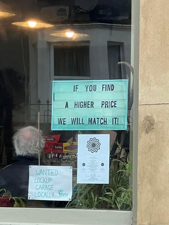 A photograph of a store window displaying two prominent signs. The top sign is a turquoise marquee-style sign reading 'IF YOU FIND A HIGHER PRICE WE WILL MATCH IT!' Below it, a handwritten white sign with green text states 'WANTED LOCKUP GARAGE LOCALLY $$$!!'. Inside the store, an elderly person with white hair is visible sitting among various items, including keychains and potted plants. The juxtaposition of the price-matching sign (typically used to promote retail goods) with the unrelated wanted ad for a garage creates unintended irony, as the two messages lack logical connection.