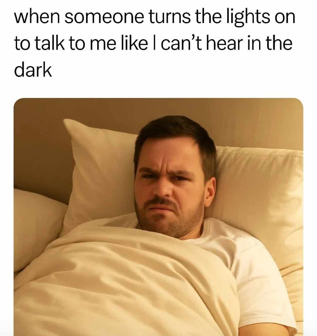This meme consists of a top text caption paired with a photograph of a man lying in bed under light-colored blankets, propped against pillows. The man has a visibly annoyed, squinting, frustrated expression, as if he was just resting or sleeping in the dark and was disrupted by bright light. The caption reads 'when someone turns the lights on to talk to me like I can’t hear in the dark', conveying a relatable, humorous feeling of irritation at the unnecessary action of turning on lights to speak to someone who is already comfortable in the dark, as hearing is not affected by lack of light.