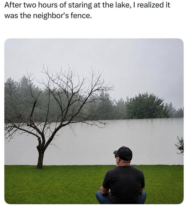 This is a humorous meme based on a real optical illusion. The text at the top reads 'After two hours of staring at the lake, I realized it was the neighbor's fence.' The photo below shows a man wearing a black cap, black t-shirt, and sunglasses, sitting on a green lawn facing what initially appears to be a calm, misty lake surrounded by trees, with a bare leafless tree on the left side of the frame. The punchline is that the supposed 'lake' is actually the flat, white surface of a neighbor's fence; the overcast, foggy weather makes the fence blend perfectly with the foggy sky background, creating the convincing illusion of a body of water. The joke plays on relatable absent-mindedness and the unexpected optical trick.