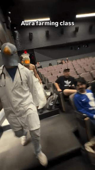 This image captures two students dressed in absurd costumes walking through the aisle of a college lecture hall, while other regular students sit in the seats watching them. The person in front wears a grey bird mask, a full white doctor's lab coat with a stethoscope, and light-colored clothing. The person behind him is shirtless, wearing a chicken mask. Overlaid text on the image reads 'Aura farming class', referencing the internet slang term 'aura farming' which describes intentionally doing bizarre, shocking things in public to build an intimidating or memorable social reputation. The joke here is that the group is literally 'farming aura' by doing this prank in the college class lecture hall.