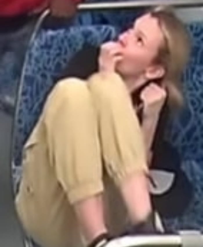 A person identified as Iryna Zarutska is sitting on a blue patterned seat, likely in a public transport setting such as a bus or train. She is wearing a yellow short-sleeved outfit and is positioned leaning back slightly, with one hand resting near her face, conveying a thoughtful or relaxed demeanor. The background includes part of another seat and a partial view of another individual in red clothing.