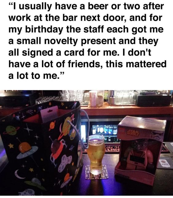 The image consists of two main parts: a top text block and a lower photograph. The text block reads: 'I usually have a beer or two after work at the bar next door, and for my birthday the staff each got me a small novelty present and they all signed a card for me. I don't have a lot of friends, this mattered a lot to me.' Below the text is a color photograph of a bar counter scene. Visible items include a black gift bag adorned with space-themed illustrations (planets, stars, a UFO), a clear glass of golden beer on a coaster, and a pink novelty item still in its cardboard packaging. The background shows typical bar elements: stacked glasses, a refrigeration unit, and bar equipment, creating a cozy, familiar atmosphere.