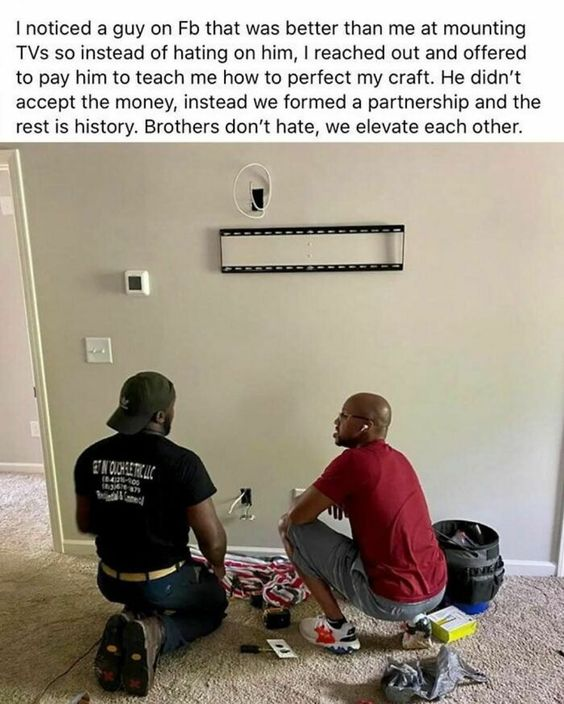 This is a wholesome, inspirational meme combining a real-life photo with a text story. The photo shows two Black men kneeling on a carpeted indoor floor, collaborating to install a TV wall mount on a light gray wall. Tools, cables, and a equipment bag are scattered around them, indicating active work on the installation. Above the image, a detailed text narrative explains that the poster noticed another man on Facebook who was more skilled at mounting TVs than himself. Instead of feeling jealous or competitive, he reached out and offered to pay the man to teach him his craft. The man declined payment, and instead, they formed a business partnership. The story concludes with the uplifting message: "Brothers don’t hate, we elevate each other." The meme emphasizes the value of collaboration over competition, uplifting fellow professionals, and building community in small business work.