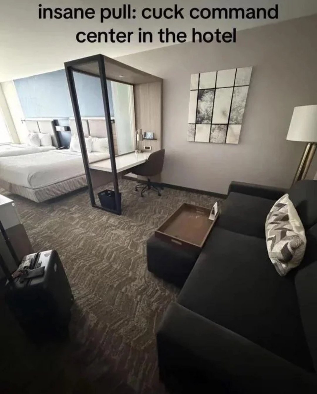 This image shows a modern hotel suite with two queen beds, a glass and metal partition separating a desk area that faces directly toward the beds, a dark couch in the foreground, patterned carpet, abstract wall art, and a rolling suitcase in the bottom left. Text overlaid at the top reads 'insane pull: cuck command center in the hotel'. The humor relies on crude internet slang: 'pull' refers to successfully inviting a sexual/romantic partner back to your accommodation, while 'cuck command center' is a vulgar, absurd joke mocking the awkward hotel layout, implying the positioned desk would be used by someone watching their partner be intimate with another person.