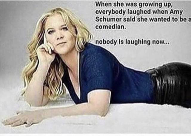 The image depicts Amy Schumer lying on a light-colored surface, wearing a dark blue top and black pants. Her left hand is positioned under her chin, and she has long, wavy blonde hair. To the right of her figure, there is white text that reads: 'When she was growing up, everybody laughed when Amy Schumer said she wanted to be a comedian. nobody is laughing now...' The text appears to critique Schumer's career by suggesting that while people initially mocked her ambition to become a comedian, her current work fails to elicit laughter, implying her comedy is unfunny.