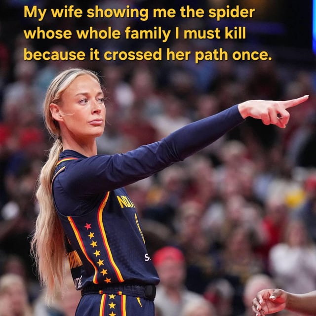 This is a relatable relationship reaction meme. The visual shows a blonde female WNBA player (Lexie Hull of the Indiana Fever) in her team uniform with a stern, serious expression, pointing firmly with her right index finger off-camera to the right, against a blurred background of a sports arena audience. The overlaid yellow text at the top of the image reads: 'My wife showing me the spider whose whole family I must kill because it crossed her path once.' The joke relies on the common relatable dynamic in many heterosexual relationships where the male partner is expected to get rid of any spiders found in the home, with the wife's demanding, no-nonsense pointing gesture exaggeratedly paired with the over-the-top requirement to kill the entire spider family just because one spider was seen.