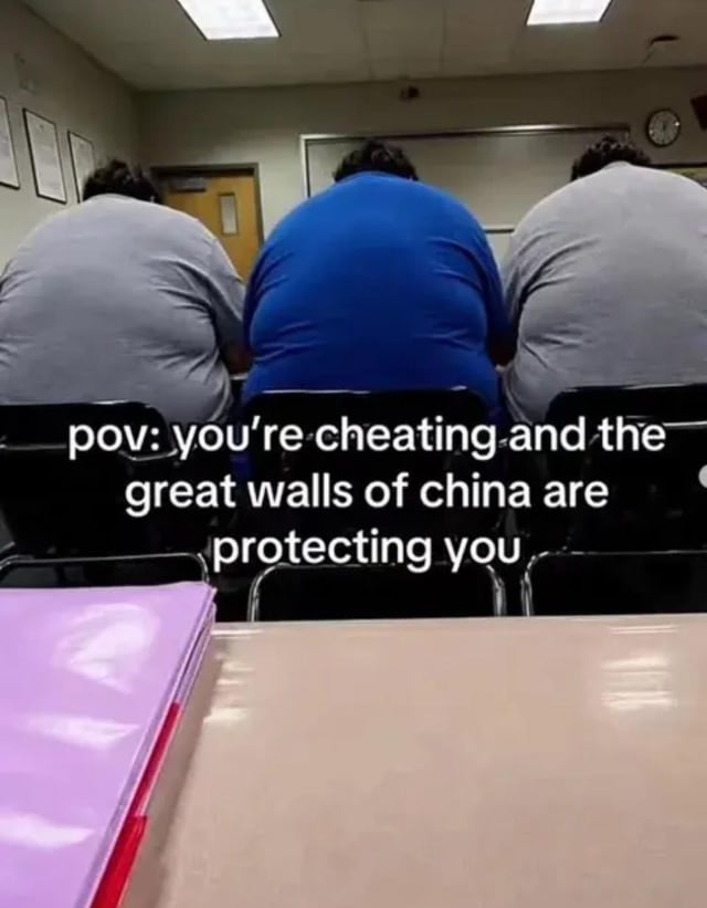 This is a first-person POV meme shot from a student's desk in a standard classroom. Three very large, broad-backed male students are seated in a row directly in front of the viewer, fully obstructing the view of anyone behind them from seeing the viewer's desk space. Overlaid text makes an exaggerated joke comparing the students' large, view-blocking backs to the Great Wall of China, which protects the viewer if they are cheating on a test or class assignment. A purple folder sits on the desk in the foreground, while standard classroom features including a whiteboard, wall clock, ceiling fluorescent lights, framed documents on the wall, and a classroom door are visible in the background.