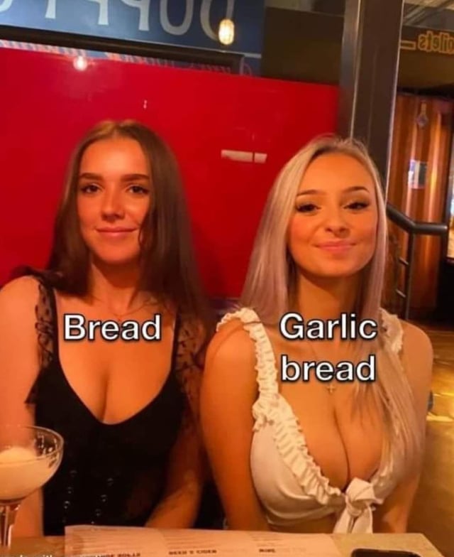This meme uses a photograph of two real women sitting side-by-side in a restaurant/bar environment to make a humorous comparison between plain bread and garlic bread. The woman on the left, labeled **Bread**, has dark hair and wears a black lace-trimmed top, smiling gently at the camera. The woman on the right, labeled **Garlic Bread**, has light blonde/silver hair and wears a white ruffled top, also smiling warmly. The joke leans into the common culinary preference for garlic bread over plain bread, framing the visually distinct (and often considered more 'appealing') woman as the 'superior' garlic bread, exaggerating the food preference into a lighthearted, playful visual gag.