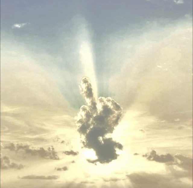 The image captures a daytime sky with soft golden-toned clouds and pale blue patches, with bright sunbeams radiating from behind the cloud cover. The central cloud formation is visually identical to a raised middle finger, illuminated by the sun rays behind it to look like a divine, heavenly gesture. The visual gag comes from the contrast between the sacred, angelic appearance of the sunlit sky and the rude, obscene gesture the cloud forms, creating an edgy, humorous effect.