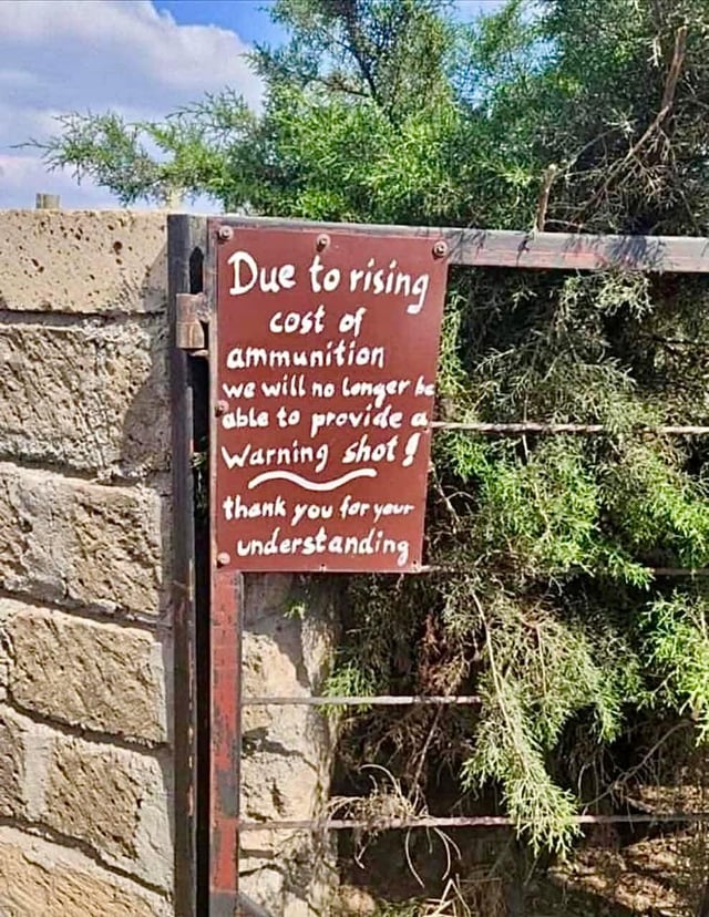 The image is a photograph of a rust-colored metal sign mounted on a metal fence positioned next to a rough stone wall, with dense green shrubbery visible behind the fence under a partly cloudy sky. The sign features handwritten white text that jokes that due to rising ammunition costs, the property owner will no longer fire warning shots at trespassers, thanking people for their understanding. The joke juxtaposes formal, polite corporate cost-cutting announcement language with the violent implication that trespassers will be shot without warning, making light of widespread inflation and rising costs applied to a private property security context.