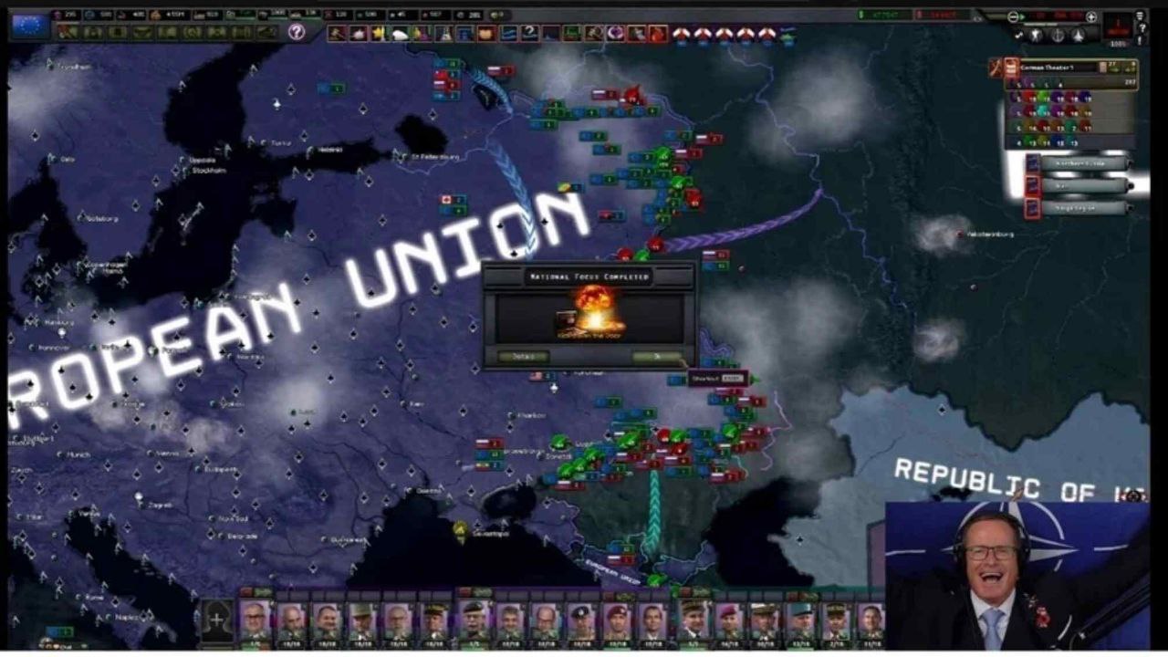 A screenshot from a grand strategy game (likely Hearts of Iron IV) depicting a map of Europe with the text 'EUROPEAN UNION' prominently overlaid. The map is filled with colored military unit icons (red, green, blue) indicating ongoing conflicts. A pop-up window labeled 'NATIONAL FOCUS COMPLETED' shows an explosion graphic. In the bottom right corner, there is an inset image of a person in a suit and tie, wearing a headset, laughing in front of a NATO flag backdrop. The combination suggests a satirical take on European Union politics and NATO involvement.
