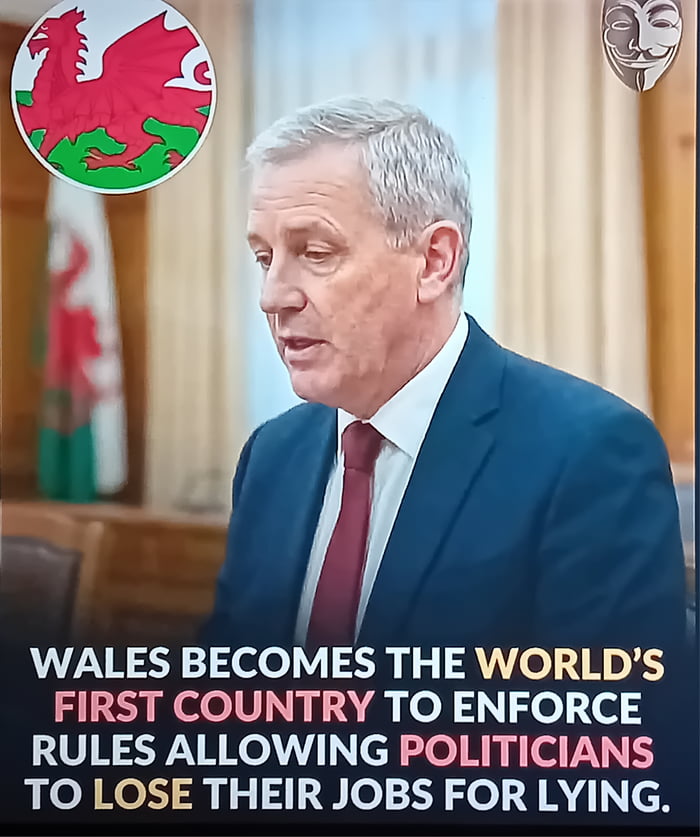 A screenshot featuring a middle-aged man in a blue suit and red tie standing in front of a background with the Welsh flag (red dragon on green and white). Overlaid text in bold, uppercase letters states: 'WALES BECOMES THE WORLD’S FIRST COUNTRY TO ENFORCE RULES ALLOWING POLITICIANS TO LOSE THEIR JOBS FOR LYING.' The setting appears to be a formal indoor environment, likely a government office or press conference, with curtains and wooden furniture visible.